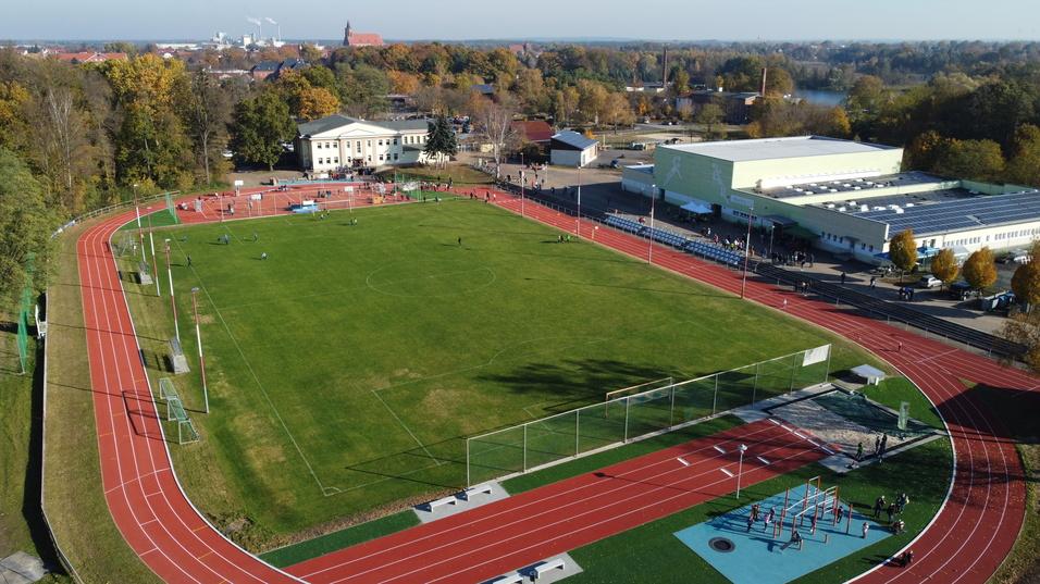 Luftaufnahme eines Sportgeländes mit Laufbahn, Fußballfeld und Spielgeräten, umgeben von Bäumen und Gebäuden.