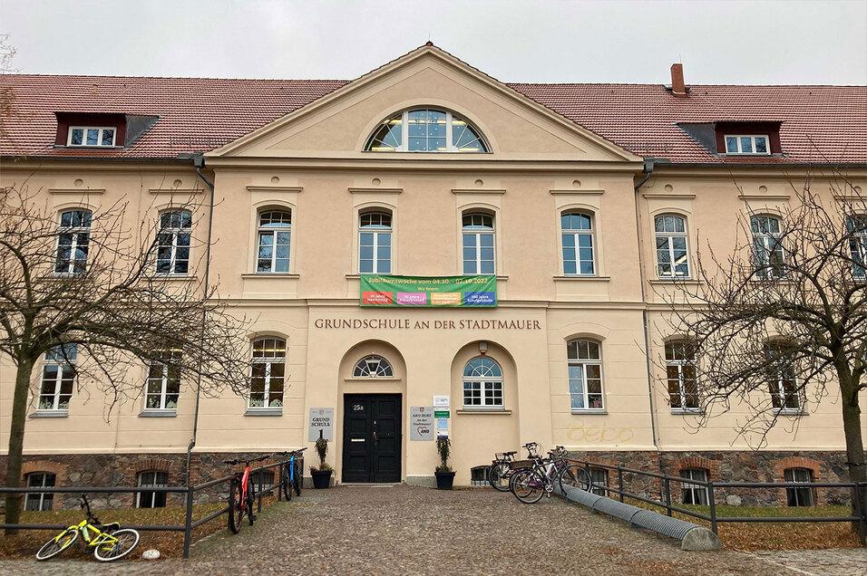 Fassade der Grundschule an der Stadtmauer mit Eingang, Banner und Fahrrädern im Vordergrund. Herbstliche Baumstämme sichtbar.