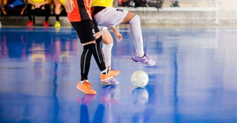 Zwei Spieler stehen auf einem blauen Hallenboden und spielen mit einem Fu&szlig;ball zwischen ihren F&uuml;&szlig;en.