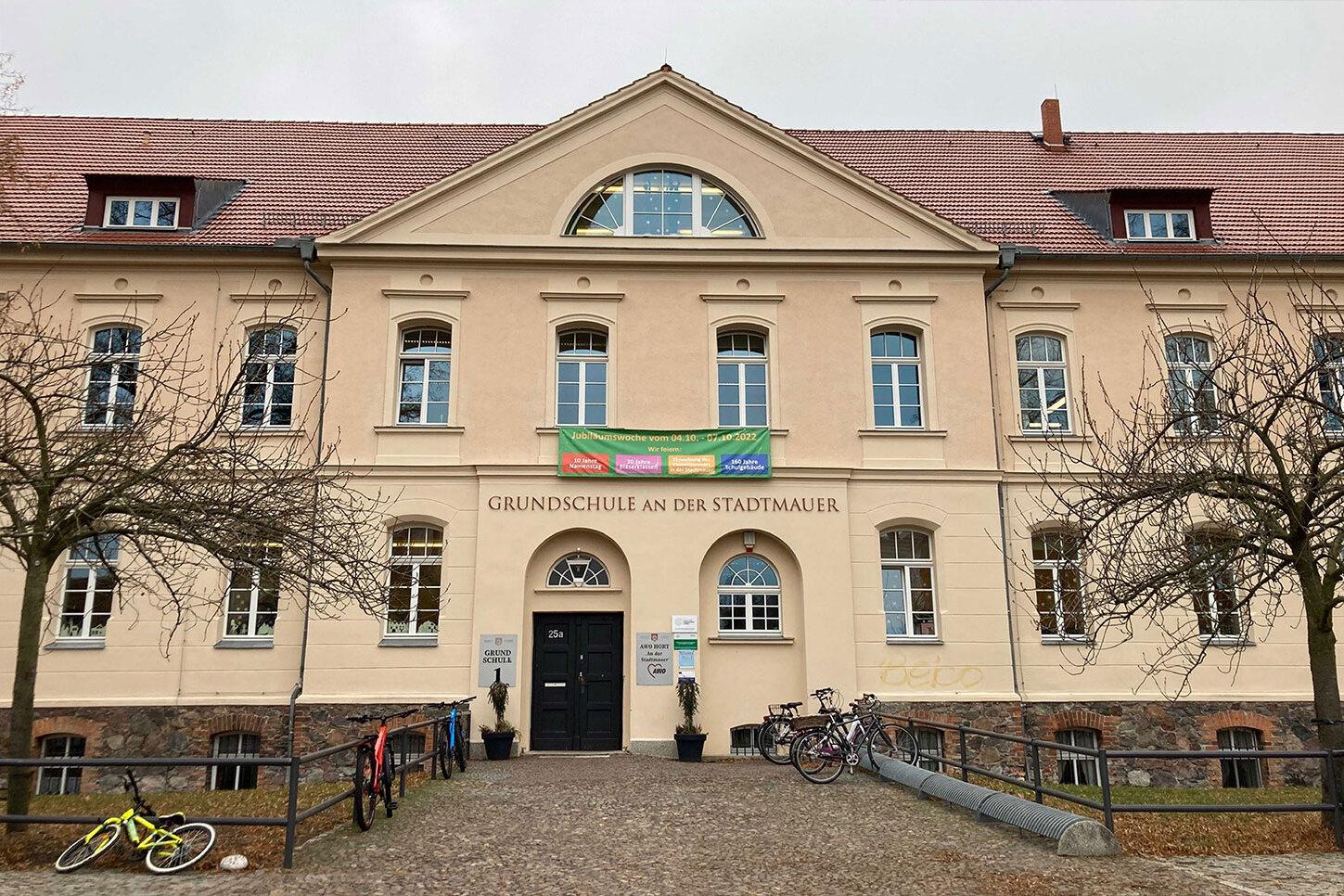 Fassade der Grundschule an der Stadtmauer mit Eingang, Banner und Fahrr&auml;dern im Vordergrund. Herbstliche Baumst&auml;mme sichtbar.