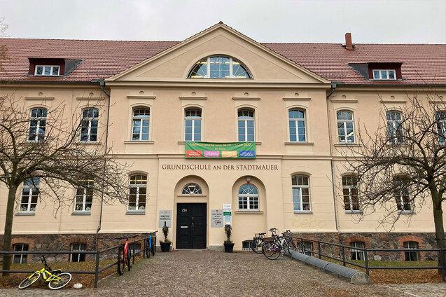 Fassade der Grundschule an der Stadtmauer mit Eingang, Banner und Fahrr&auml;dern im Vordergrund. Herbstliche Baumst&auml;mme sichtbar.