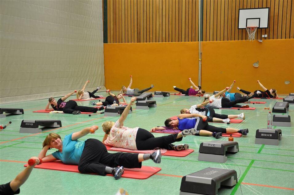 Gruppe von Frauen macht Übungen auf Matten in einer Sporthalle, einige nutzen Hanteln und Fitnessblock.