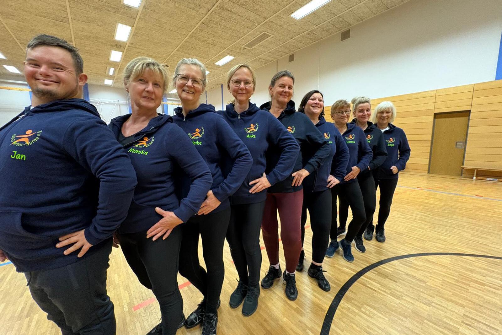 Gruppe von neun Personen in blauen Sweatshirts, die in einer Sporthalle aufgestellt stehen und in die Kamera lächeln.