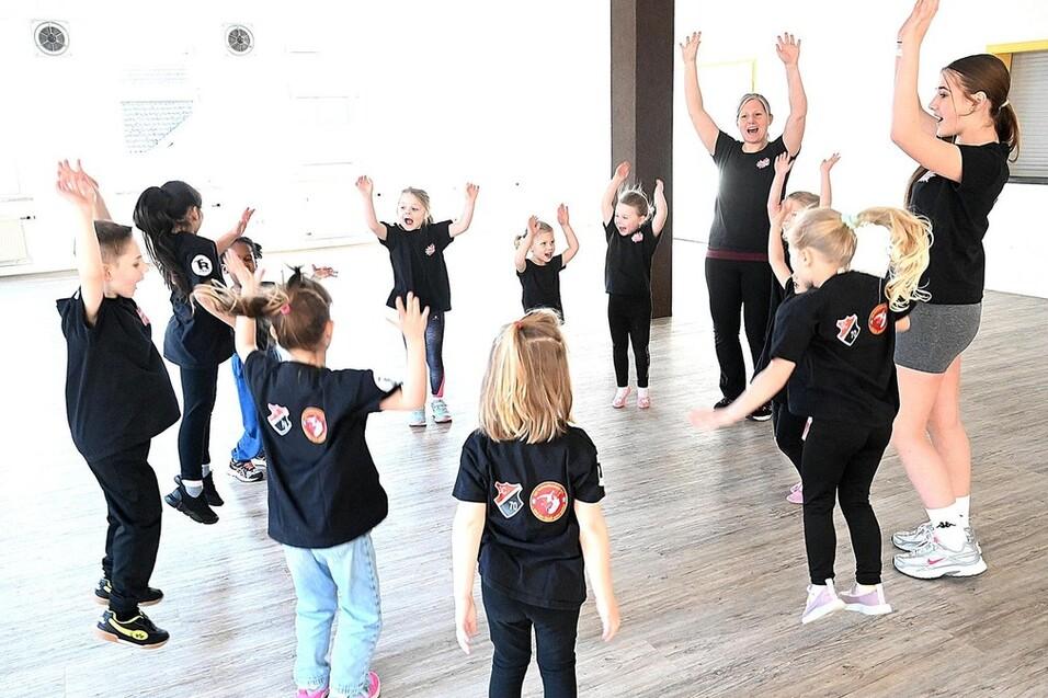 Gruppe von Kindern in Schwarz, die in einem hellen Raum mit erhobenen Armen zusammen tanzen und Freude zeigen.