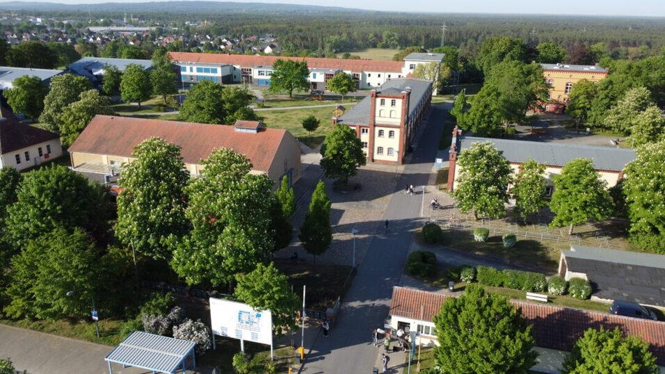 Luftaufnahme eines Schulgeländes mit mehreren Gebäuden und grünen Bäumen auf einem sonnigen Tag.
