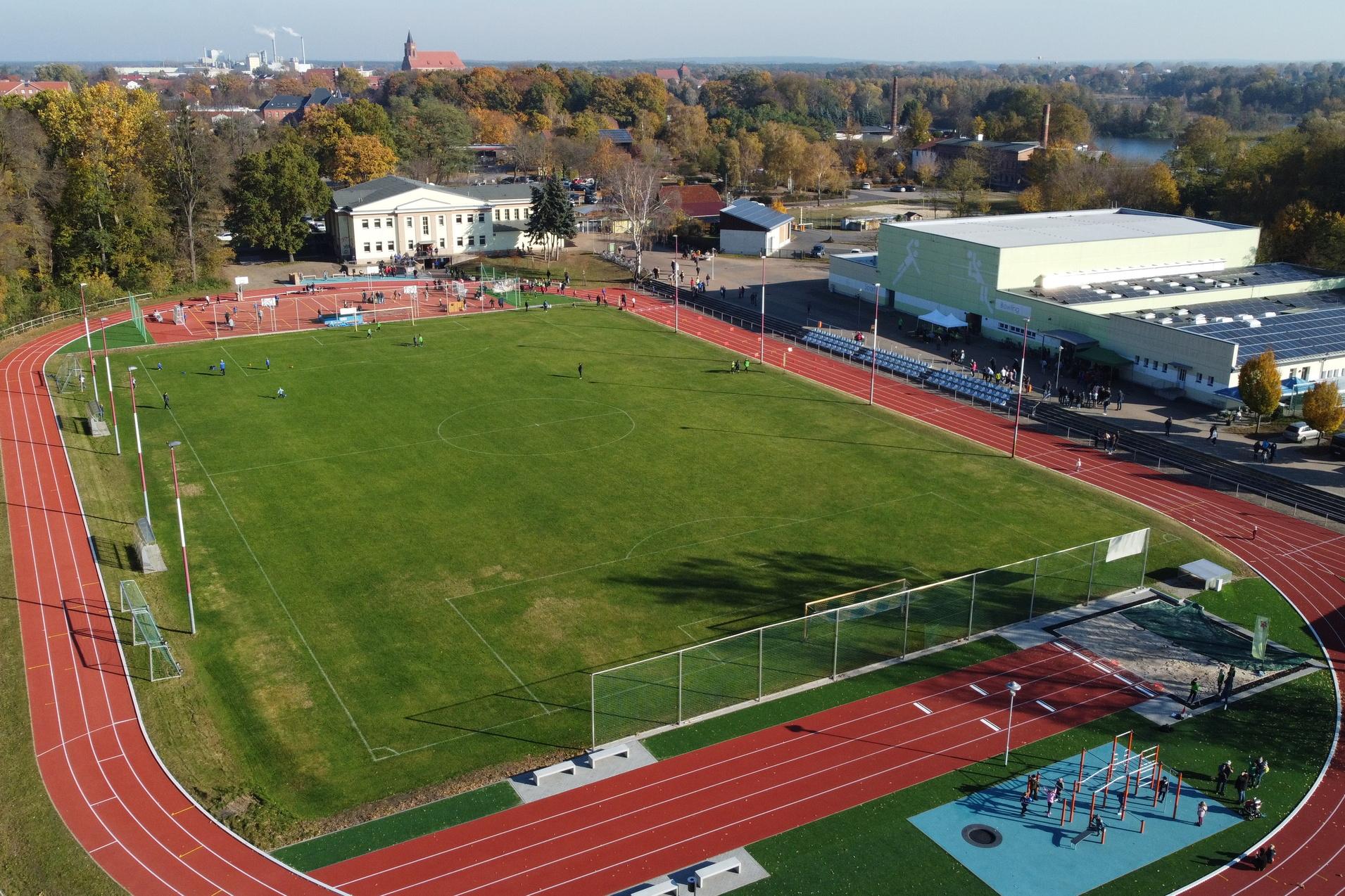 Luftaufnahme eines Sportgel&auml;ndes mit Fu&szlig;ballplatz, Laufbahn und Freizeitanlage. B&auml;ume und Geb&auml;ude im Hintergrund.