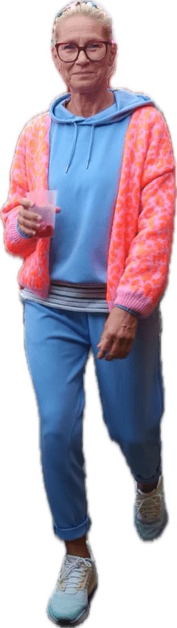 Frau mit Brille in rosa-weißer Strickjacke und blauen Kleidungsstücken, hält einen Becher und geht.