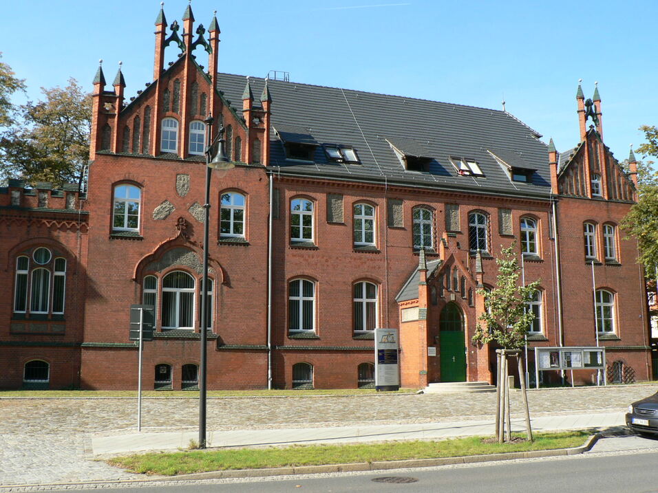 Historisches rotes Ziegelgeb&auml;ude mit Spitzdach, Fenstern und T&uuml;r im neugotischen Stil, umgeben von B&auml;umen.