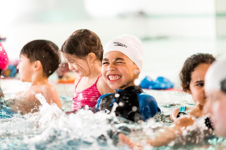 Junge im Schwimmbecken mit weißer Badekappe lächelt, umgeben von weiteren Kindern und spritzendem Wasser.